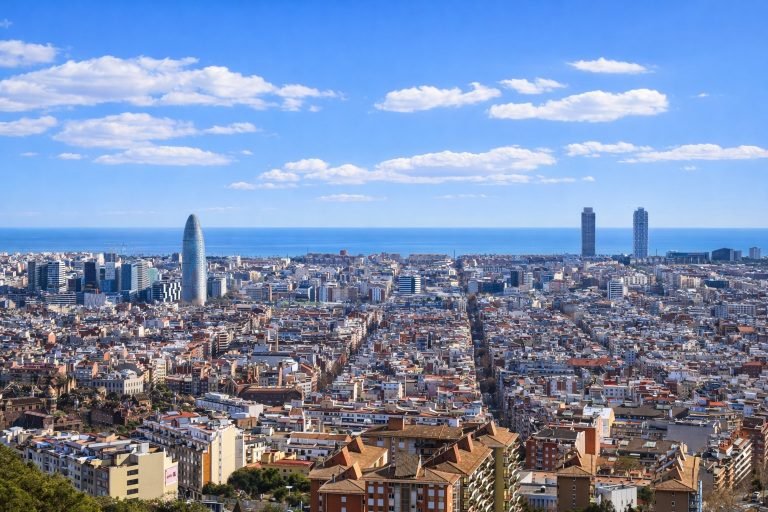 Panoramic cityscape of Barcelona stretching to the Mediterranean Sea, with Torre Glòries and coastal high-rises under a bright blue sky.