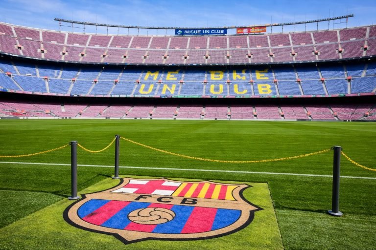 Wide panoramic view inside Camp Nou stadium in Barcelona with the FC Barcelona crest on the pitch and “Més que un club” displayed in the stands on a sunny day Caption Description