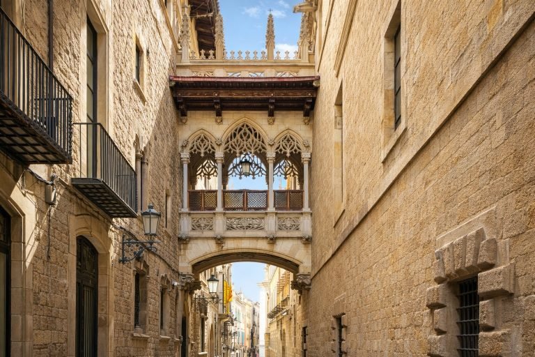 Pont del Bisbe in Barcelona’s Gothic Quarter on a bright sunny afternoon, showing the ornate neo-Gothic bridge spanning a narrow stone street beneath blue skies.