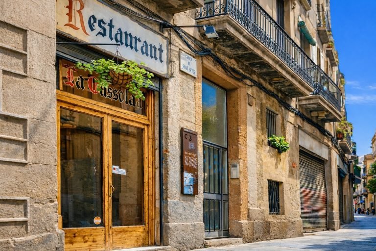 Things to do in Barcelona - Sunlit narrow street in Barcelona with traditional stone buildings, balconies, and the historic Restaurant La Cassola facade