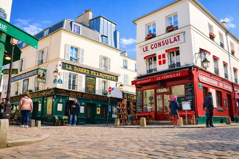 Things to do in Paris - Sunny Paris café corner with Le Consulat and La Bonne Franquette in Montmartre, cobblestone street, outdoor seating, and blue skies.