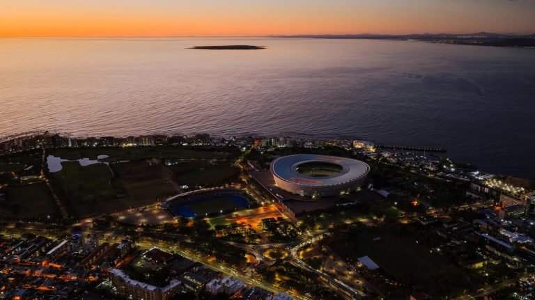 Aerial sunset view of Cape Town’s coastline with Cape Town Stadium lit up near the waterfront and the ocean stretching to the horizon.