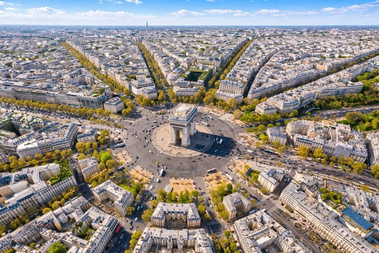 Aerial view of the Arc de Triomphe at Place Charles de Gaulle in Paris on a bright, sunny afternoon, with radiating avenues, Haussmann buildings, and clear blue skies.