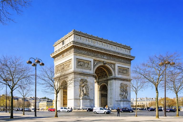 The Arc de Triomphe in Paris on a bright, sunny afternoon, shown in a wide landscape view beneath clear blue skies.