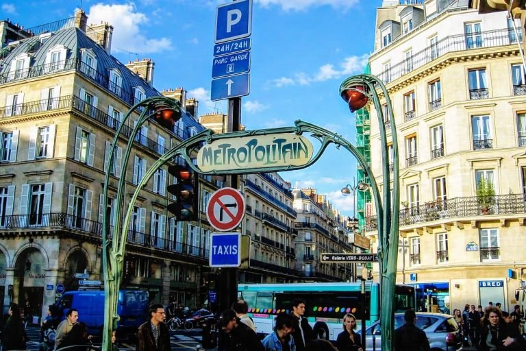 Art Nouveau Métropolitain entrance to the Paris Metro on a bright, sunny afternoon, with pedestrians, Haussmann-style buildings, and clear blue skies. Caption: Description: