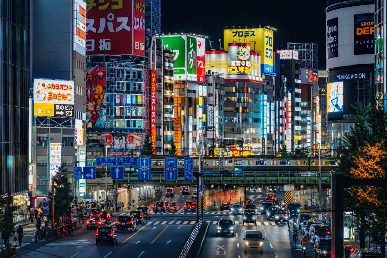 Night view of central Tokyo with neon-lit billboards, busy streets, and traffic flowing beneath elevated rail tracks.