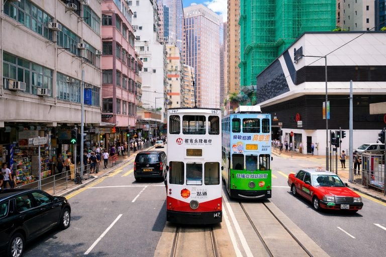Double-decker trams and taxis on a busy Hong Kong street, with sharp details, vivid colors, and bright sunlight under clear blue skies.