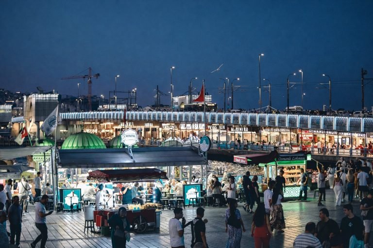 Evening street food scene near Galata Bridge in Istanbul, with crisp lights, sharp details, and crowds gathered around illuminated food stalls by the water. Caption Description