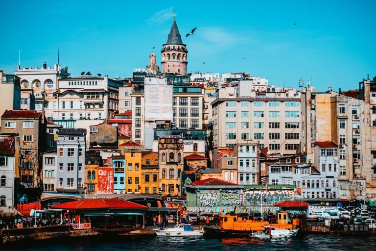 Galata Tower rising above colorful waterfront buildings in Istanbul, with crisp architectural details, bright façades, and boats along the Golden Horn under a clear blue sky.