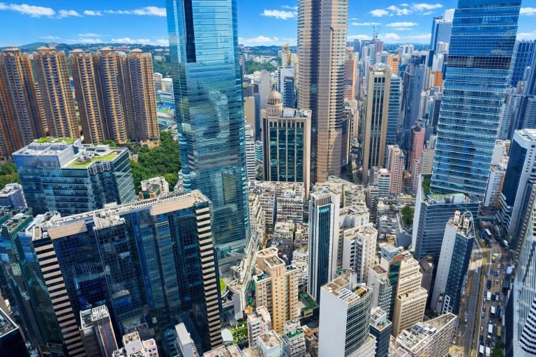 Aerial view of Hong Kong’s dense skyline with glass skyscrapers and tightly packed buildings under bright blue skies on a sunny day.