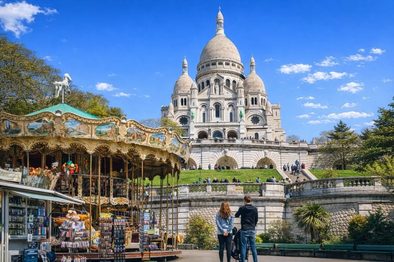 Sacré-Cœur Basilica in Montmartre on a bright, sunny afternoon, with a colorful carousel in the foreground and clear blue skies above Paris.