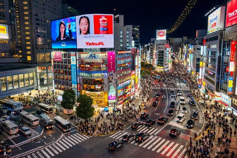 Shibuya Crossing in Tokyo at night with bright neon billboards, clear zebra crossings, and heavy pedestrian and car traffic.