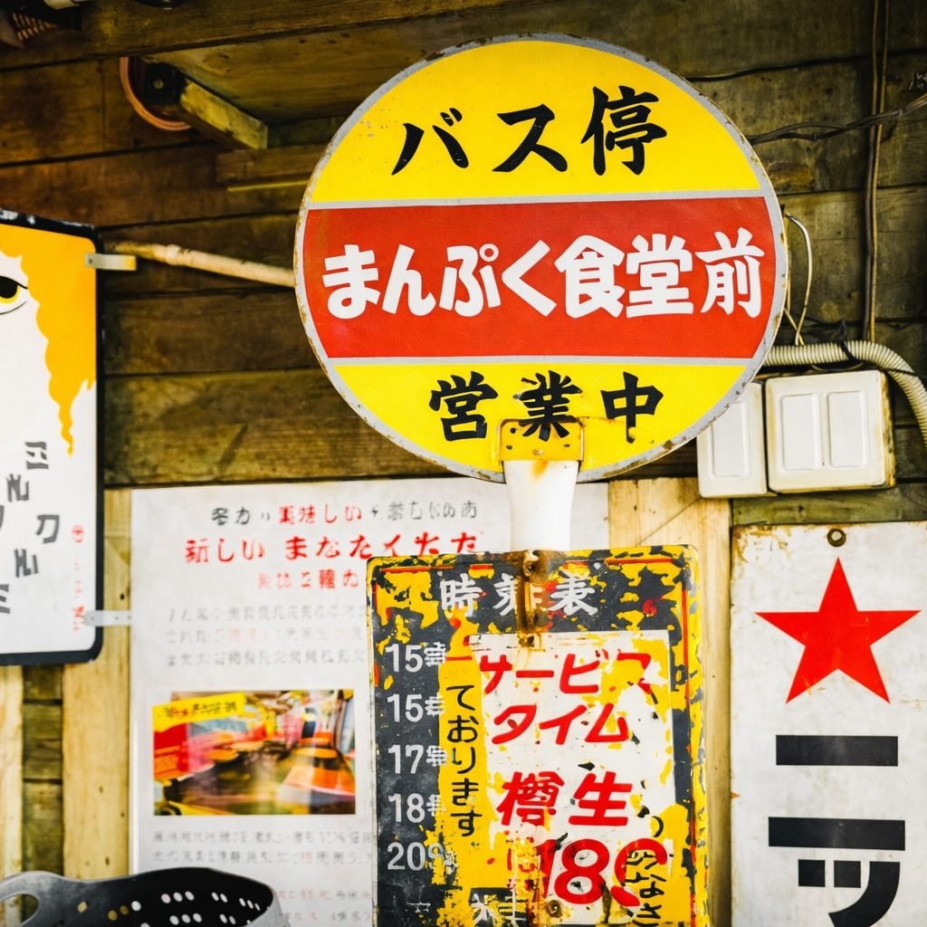 Japanese street sign in landscape format showing a bus stop marker and local eatery signage with handwritten notices and colorful posters.