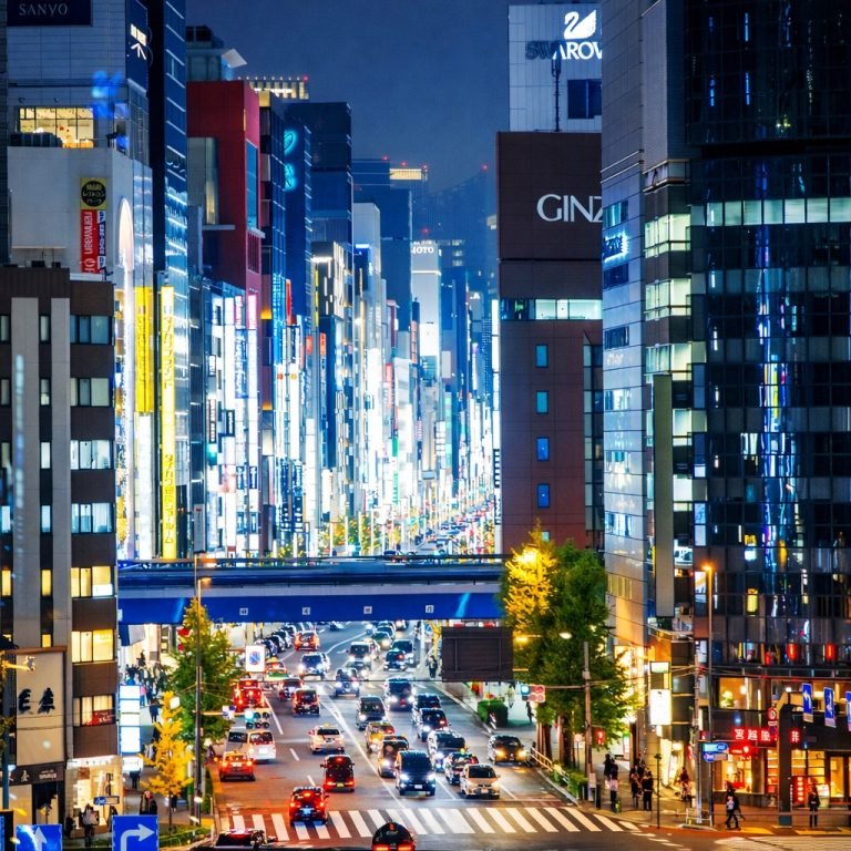 Bright nighttime view of a busy Tokyo street with illuminated buildings, heavy traffic, and neon signage stretching into the distance.