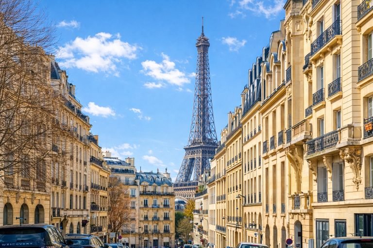 Sunny Paris street framed by Haussmann buildings with the Eiffel Tower rising in the distance under clear blue skies.