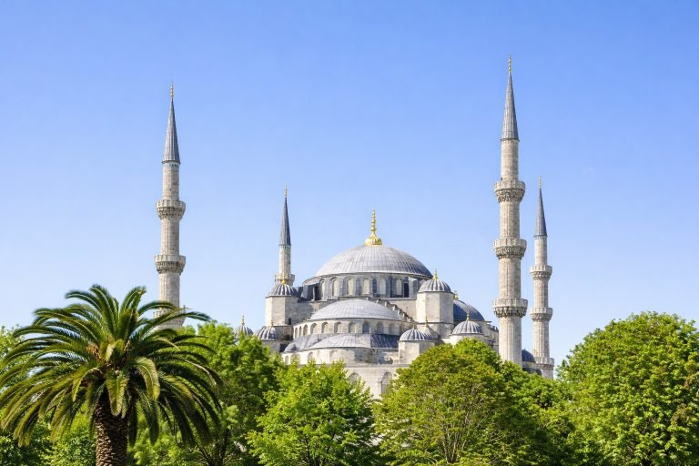 Blue Mosque (Sultan Ahmed Mosque) in Istanbul under a clear blue sky, with crisp stone details, six minarets, and lush green trees in the foreground.