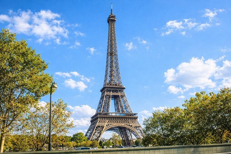 The Eiffel Tower in Paris on a bright, sunny afternoon, framed by trees and a clear blue sky in a wide landscape view.