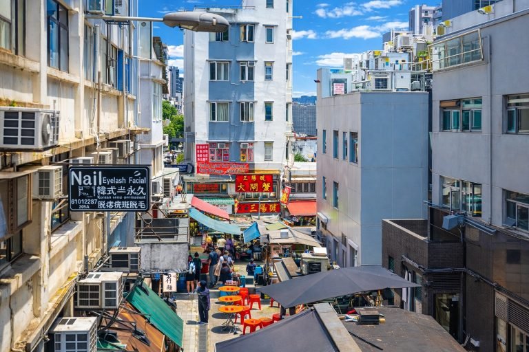 Street-level view of a narrow Hong Kong alley with market stalls and shop signs, shown in bright sunlight with vivid colors and a clear blue sky. Caption: Description: