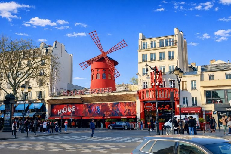 Moulin Rouge cabaret in Paris with its iconic red windmill on a bright sunny afternoon under clear blue skies, surrounded by lively street activity. Caption: Description: