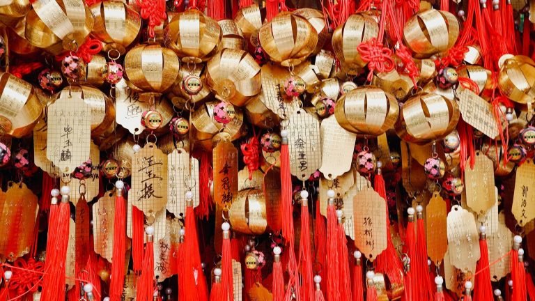 Close-up of traditional Chinese gold lantern ornaments and prayer tags with red tassels hanging closely together. Caption: Description: