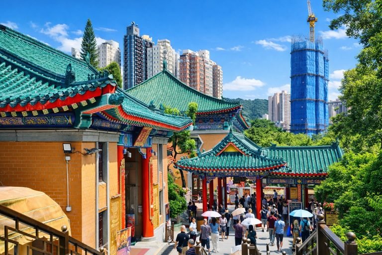 Traditional Chinese temple in Hong Kong with green tiled roofs and red pillars, surrounded by modern high-rise buildings on a bright sunny day with blue skies.