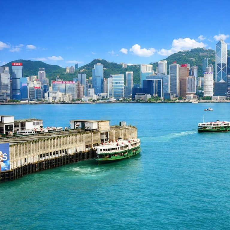 Bright daytime view of Victoria Harbour with Star Ferry boats crossing turquoise water and Hong Kong’s skyline under clear blue skies. Caption: Description:
