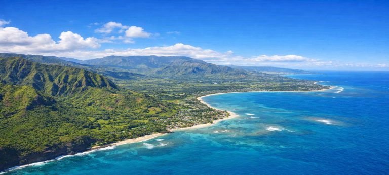 Aerial view of Kauaʻi showing lush green mountains, rugged coastline, turquoise ocean waters, and sandy beaches under clear blue skies on a sunny afternoon Caption: Description: