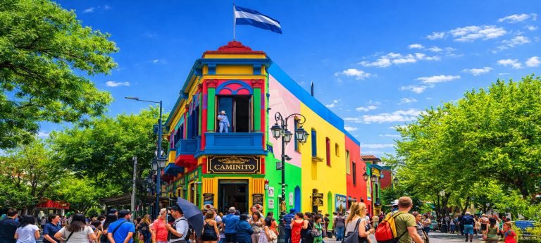 A vibrant, multi-colored corner building in the Caminito street museum of La Boca, Buenos Aires, featuring bright yellow, blue, and red facades under a clear blue sky. A crowd of tourists walks along the street in front of the building, which is topped with an Argentine flag.