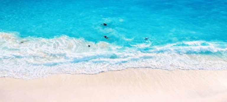 Aerial view of swimmers in crystal-clear turquoise water as gentle waves roll onto a tropical white sand beach under bright afternoon sun.