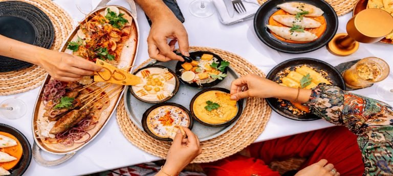 Overhead view of friends sharing a colorful spread of Tanzanian-style dishes with dips, grilled skewers, seafood, and fresh herbs on a bright white table.