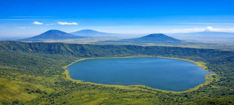 Aerial view of a lush volcanic crater with a deep blue lake at the center, surrounded by green forested slopes under a bright blue sky.