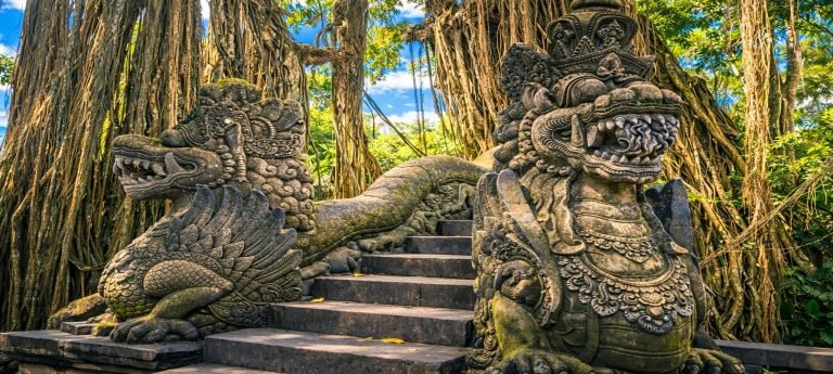 Stone guardian statues lining a stairway beneath towering banyan trees in the Sacred Monkey Forest Sanctuary, Ubud, Bali, on a bright sunny day.