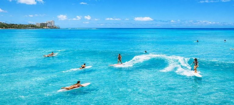 Surfers riding small waves in clear turquoise water off a Hawaiian beach on a sunny day, with calm ocean conditions and light clouds overhead