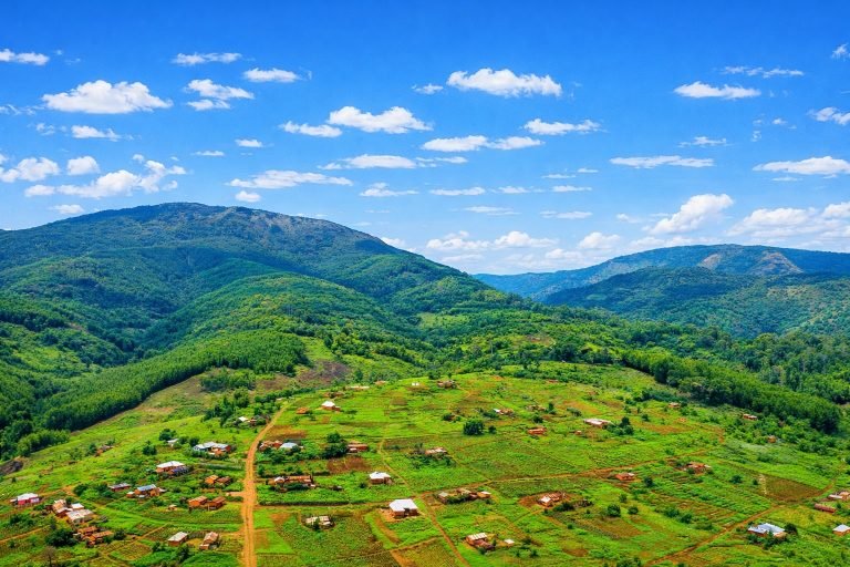 Sunlit green mountain valley with scattered rural homes, farmland plots, and rolling forested hills beneath a bright blue sky. Caption: Description: