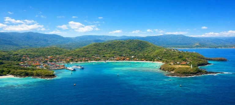 Aerial view of a tropical bay in Bali with turquoise water, a coastal village, boats in the harbor, and green hills under a bright blue sky.