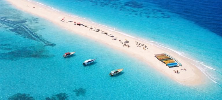 Aerial view of a narrow white sandbar in Zanzibar surrounded by clear turquoise ocean with four boats anchored nearby.