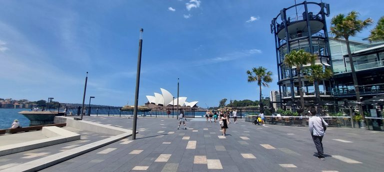 Things to do in Sydney Australia: View of the Sydney Opera House from a waterfront promenade on a sunny day with people walking and blue skies overhead.