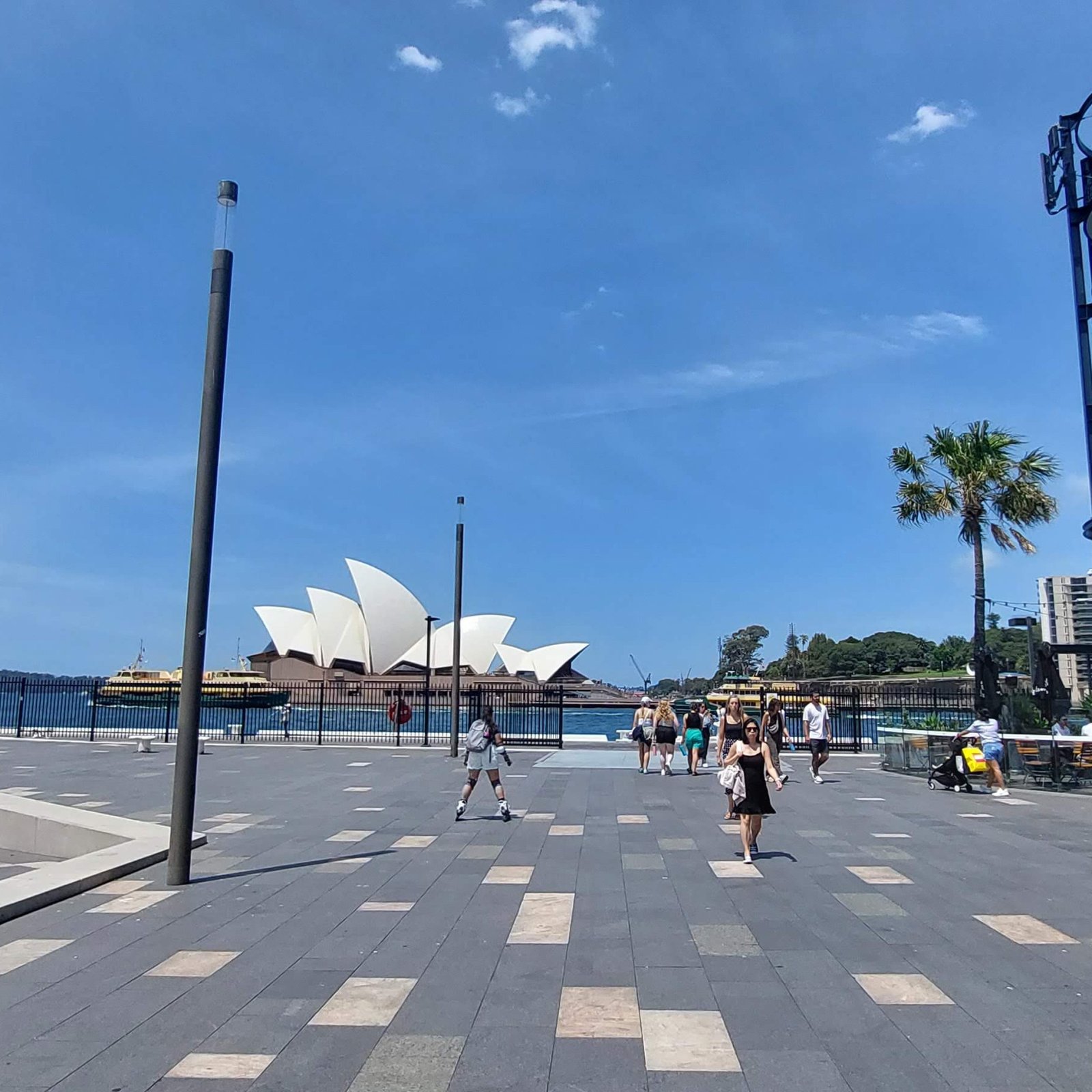 Things to do in Sydney Australia: View of the Sydney Opera House from a waterfront promenade on a sunny day with people walking and blue skies overhead.