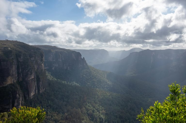 Day Trips from Sydney: Sunlit mountain valley and cliff formations with misty forest layers, Blue Mountains near Sydney, Australia.