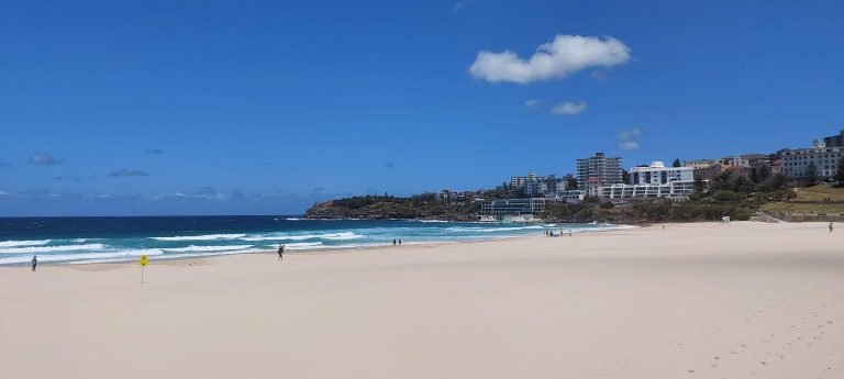 Sunny daytime view of Bondi Beach in Sydney with wide sandy shoreline, turquoise waves, and coastal buildings under a bright blue sky.