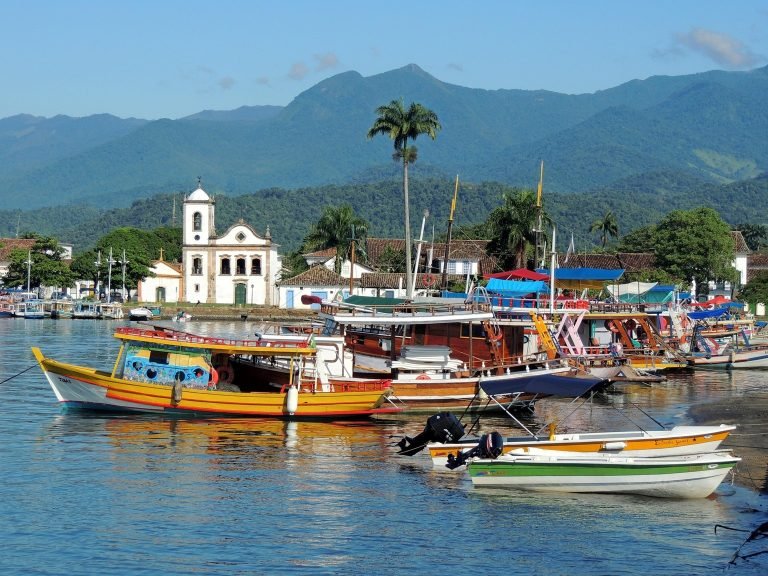 Colorful wooden boats docked in Paraty harbor with colonial church and mountains in the background in Brazil Caption Description