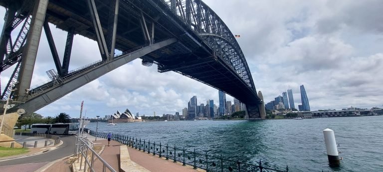 View from beneath Sydney Harbour Bridge looking across the water toward the Sydney Opera House and the city skyline.