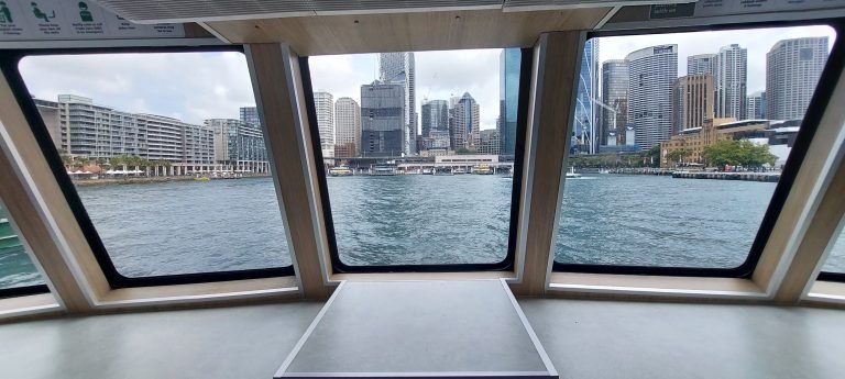 Sydney skyline viewed from inside a ferry through a large window, with harbour water in the foreground.