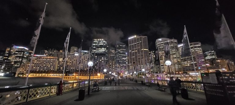 Things to do in Sydney Australia: Night view of Sydney Harbour with a pedestrian walkway, city skyscrapers, and lights reflecting in the distance.