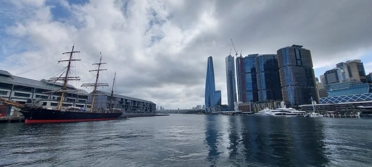 Barangaroo waterfront in Sydney with modern skyscrapers reflected on the harbour under a cloudy sky.