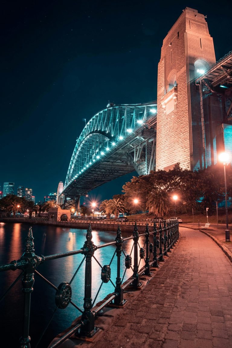 Sydney Harbour Bridge at night, lit in blue with reflections on the water along the harbour walkway.