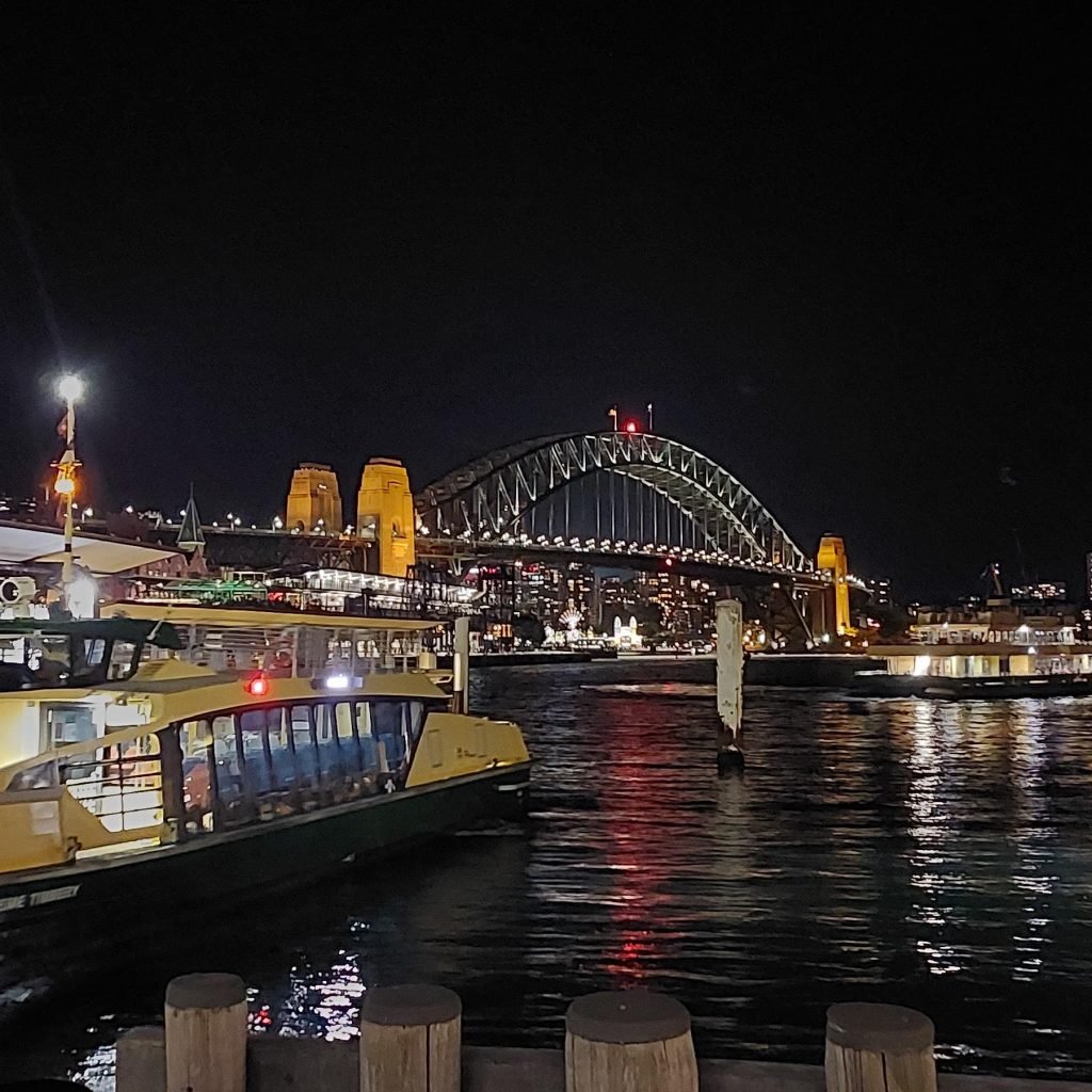 Sydney Harbour Bridge illuminated at night, viewed from Circular Quay with a ferry in the foreground and lights reflecting on the water.