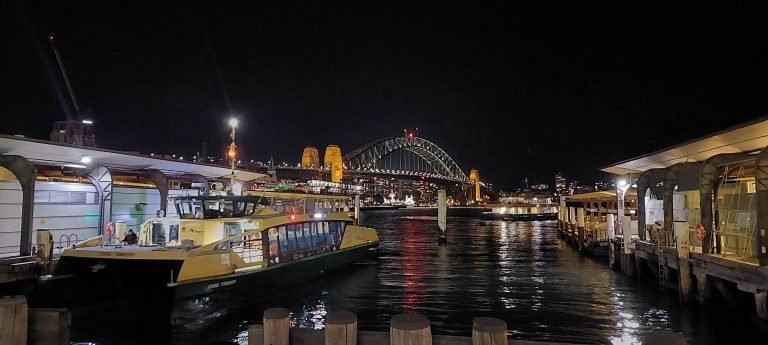 Sydney Harbour Bridge illuminated at night, viewed from Circular Quay with a ferry in the foreground and lights reflecting on the water.