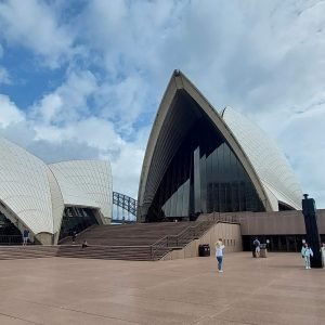 Sydney Opera House forecourt with visitors walking near the iconic sails under a partly cloudy sky.