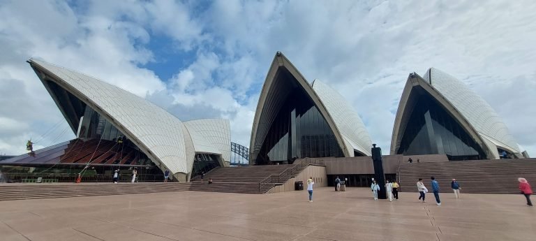 Sydney Opera House forecourt with visitors walking near the iconic sails under a partly cloudy sky.